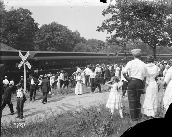 View from behind fence of large crowd of people getting off railroad cars. Men, women and children are standing on or near a dirt road near a railroad crossing sign.