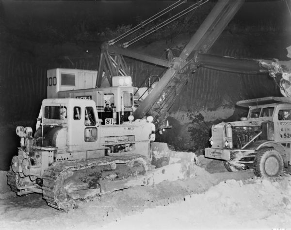 Crawler Tractor with Bucyrus Erie Crane and Dump Truck On the left is a man operating a crawler tractor, and behind him are two men in a Bucyrus Erie Crane. Another man is driving a dump truck on the right.