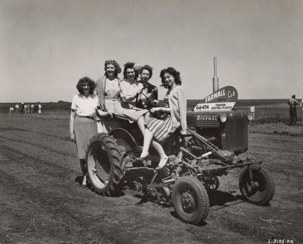 Five female Harvester employees perched on a McCormick-Deering Farmall tractor in Minneapolis, Minnesota. Taken at Farmall Cub Presentation Demonstration. A sign on the Farmall reads: "Farmall Cub with Four-Row Vegetable Planter. Cub-474." Left to right: Lois Tessum, Muriel Johnson, Faith Tessum, Maude Allen, Mae Rudell, all of the Office of Minneapolis, Minn. Branch, 