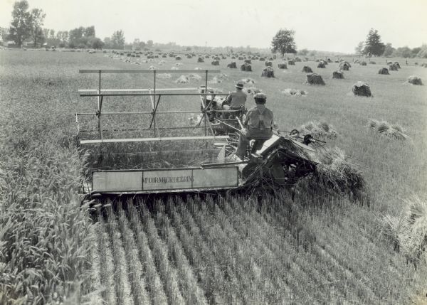Elevated rear view of two men in a field operating a new McCormick-Deering F-20 tractor and M-4 power binder, which was owned and operated by Grover Evans, R.R. 2, Kirklin, Indiana. Farm buildings are in the distance on the left.
