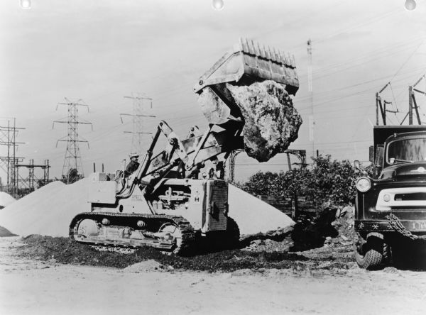 International Harvester TD-15 Crawler Tractor A man is operating the crawler tractor outdoors. The crawler is carrying a large rock towards a truck parked on the right. Power lines are in the background. Caption reads: "International Harvester TD-15 Crawler Tractor with Drott Four-In-One Attachments." Text on back of print reads: "N.M. Pierre, Skokie, Ill."