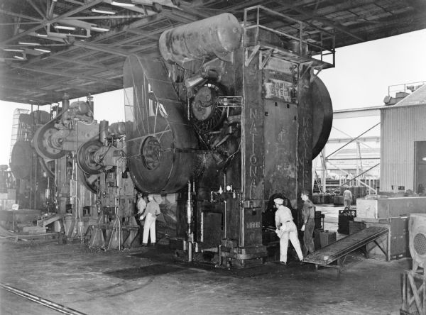 Forging Equipment with Induction Billet Heating Equipment Man are working with equipment set up underneath an opensided, roofed building.