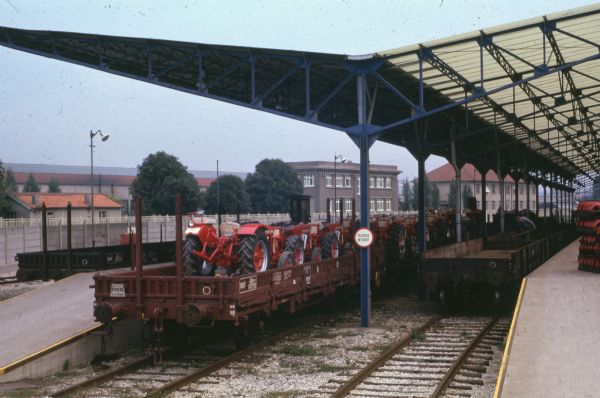 International Harvester in France Outdoor view of tractors on a railroad car near a loading dock under a roof. There is a man working on an empty railroad car in the background on the right. A sign on a post reads: "Passage Interdit."
