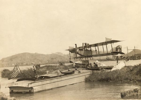 A Curtiss hydroaeroplane tied down prior to a test flight over San Diego Bay.  Two unidentified pilots rest on the dock in the shade of its wing.