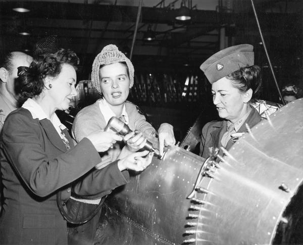 A real-life Rosie the Riveter instructs journalists on the Aviation Writers' Tour at the Goodyear Aircraft Factory. This plant manufactured Martin Marauders, FG-1 Corsairs, and lighter-than-air ships. The other writer is wearing a Civil Air Patrol civilian uniform. The tour for these and other aviation writers was organized by Goodyear publicist, Harry Bruno.