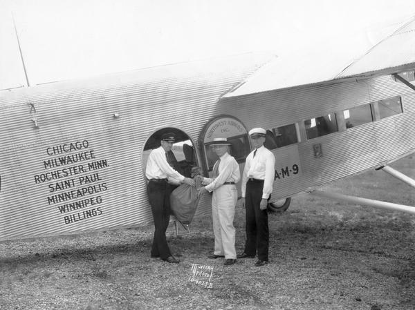A Northwest Airways crew delivers a bag of airmail to Madison postmaster W.A. Devine. With him, posed in front of the Ford TriMotor, are pilots Elmer Wahl (left) and Lee Smith. A passenger can be seen in the plane window to Smith's right.