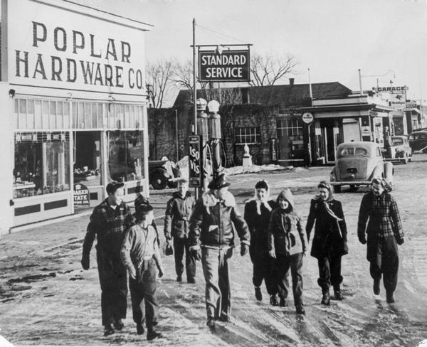 Elvated view of war hero Richard Ira Bong (center, in flight jacket) on leave in Poplar, with friends, relatives, and admirers. The group is walking down a main street on which the Poplar Hardware Company store and a Standard gasoline station can be seen.