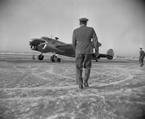 A Wisconsin Central Airlines pilot walking toward a twin-engine Lockheed 10A airplane parked on the runway at Truax Field.