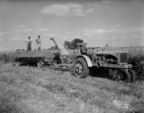 Allis Chalmers tractor pulling a chopper and a wagon, with one man driving the tractor, two men on the wagon, and one man talking to the driver.