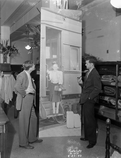 George Stein, photographer, on the right, and Frank Marx, window decorator, on the left in Manchesters Department Store. They are looking at a 10' tall poster of a female model stepping off a train.