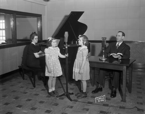 Reverend C.O. and Mrs. Grobber and two daughters at WIBU radio station studio. Reverend Grobber is sitting in front of a microphone, Mrs. Grobber is at the piano, and the daughters are standing at another microphone.