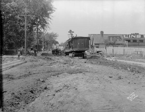 Steam shovel loading a truck as part of road construction in front of Collins Lumber Co., 2308 University Avenue. There is a horse-drawn wagon passing by. Text on back of print reads: "Univ. Ave. Construction at Walnut St. — looking W. Steam Shovel loading truck. Collins Lumber Co. -2308."