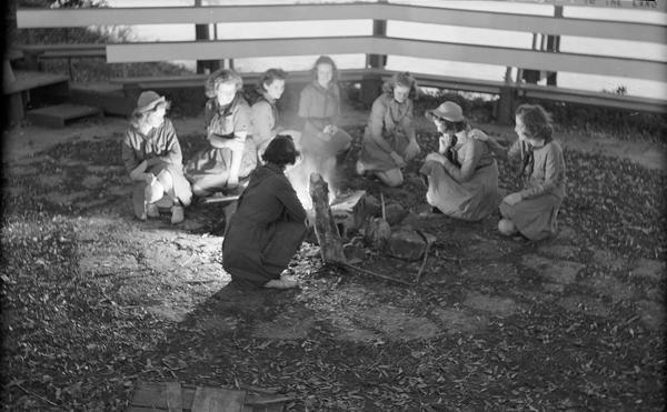 Eight Girl Scouts kneeling around a campfire inside a campfire ring at Camp Indianola.