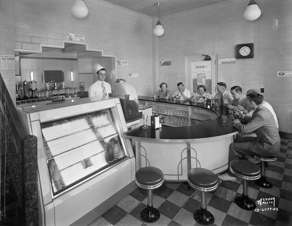 A male soda jerk is standing behind the semi-circular counter in the Bancroft Dairy fountain room, with seven customers eating ice cream.