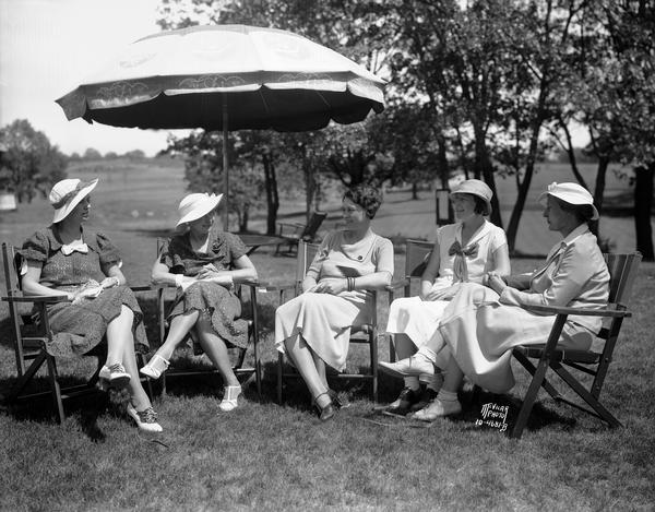 Women are relaxing under an umbrella on a sunny day on the grounds of the Nakoma Country Club. Seated left to right: Mrs. Fred Parr, Mrs. E.C. Holt, President, Mrs. C.R. Reierson, Mrs. S.M. Schoyen, and Mrs. W.H. Angell.