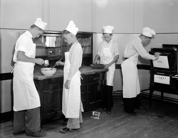 Four boys in chef's hats and aprons are preparing food at a counter and stove in the East High School boys' cooking class. Left to right: Arthur Hanson, Kenneth Elliot, Lu Verne Henry, and Benny Sherven.