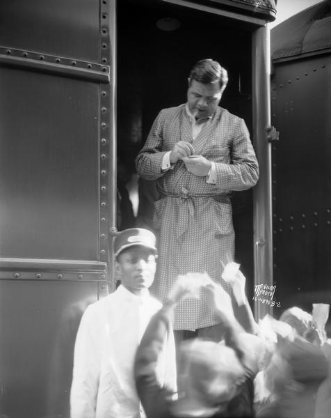 Babe Ruth, wearing dressing gown, stands in the doorway of a train and signs autographs for clamoring fans, with a train porter in uniform standing by.
