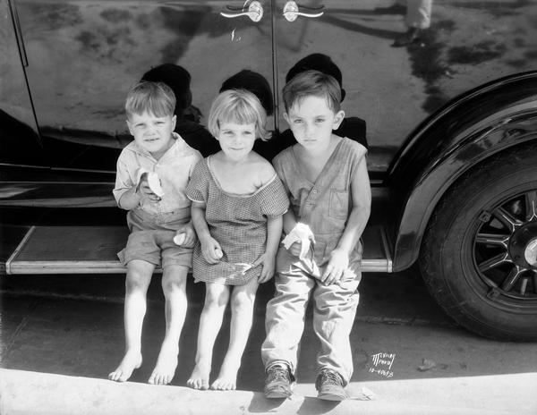 Three lost children are sitting on the running board of a car and eating bananas. Left to right: Bobbie Simpson, 5, Vina Mae Simpson, 4, Harold Ring, 4. They wandered off and were found on the east side of Madison.