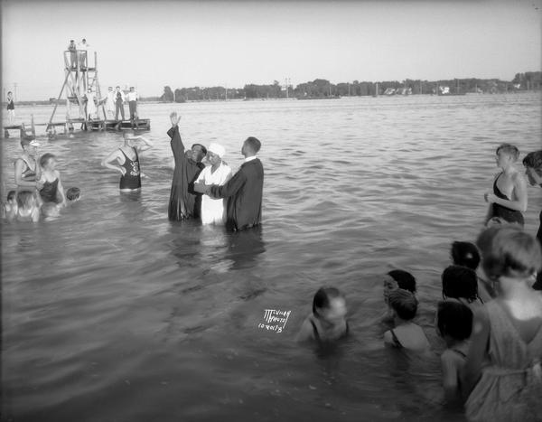 Evangelist W.P. Butler with hand upraised and Rev. Joseph Washington, pastor of Mt. Zion Baptist Church stand beside Lulu Elroy who is about to be baptized by immersion in Lake Monona. A lifeguard and swimmers are looking on.