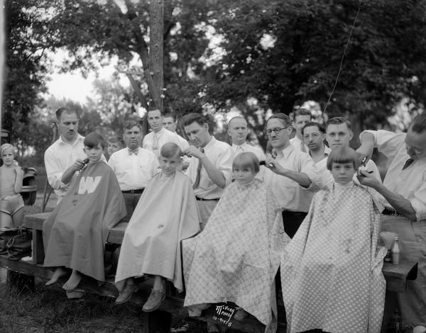 Barbers cutting the hair of four children at The Capital Times Kiddie Camp, with other barbers and children looking on.