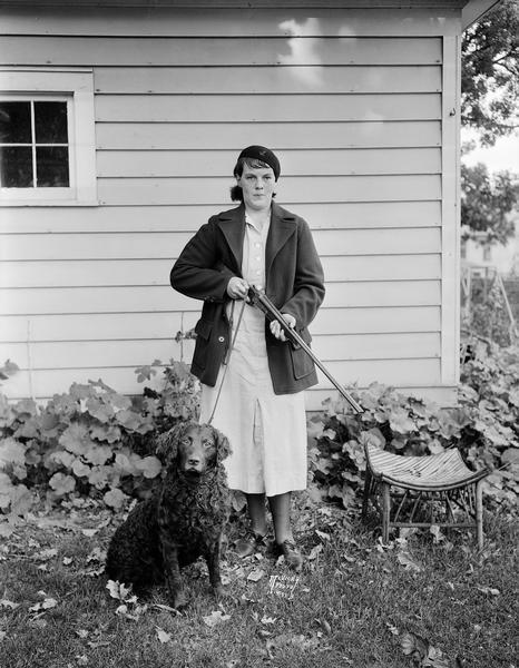 Ruth L. Brissee holding a rifle, standing with her dog in a garden at 2437 Fox Avenue. At age 15 she is the youngest huntress in Dane County.