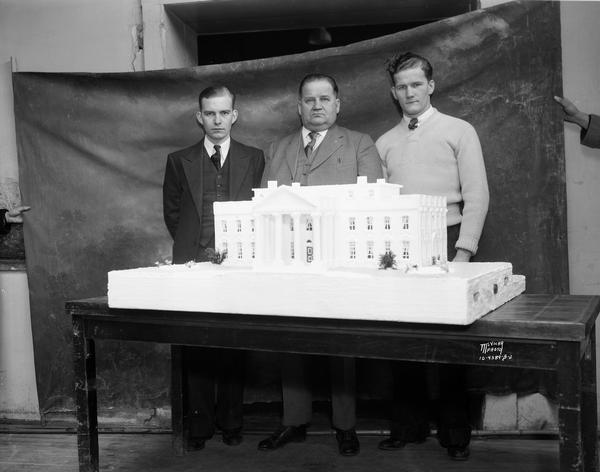 Three men are standing behind a cake shaped like the White House, baked by the Strand Baking Company, 2007 Atwood Avenue, for the Roosevelt Birthday Ball. Men are holding a backdrop behind the men.