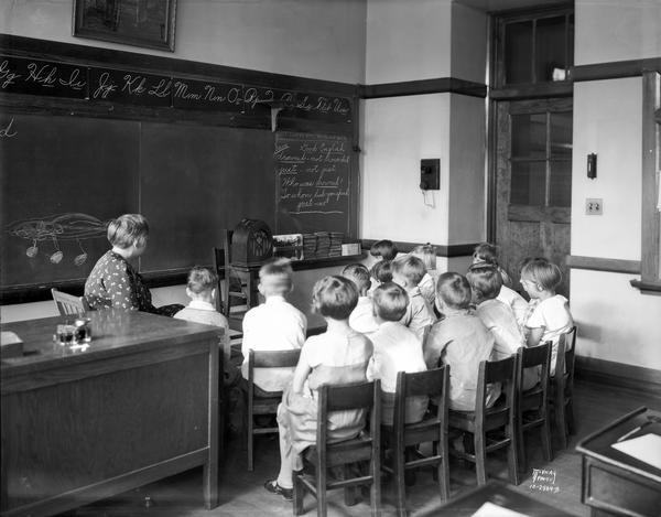 Emerson School children and teacher listen to a radio broadcast from a radio set up in front of the blackboard in the classroom at 2421 East Johnson Street.