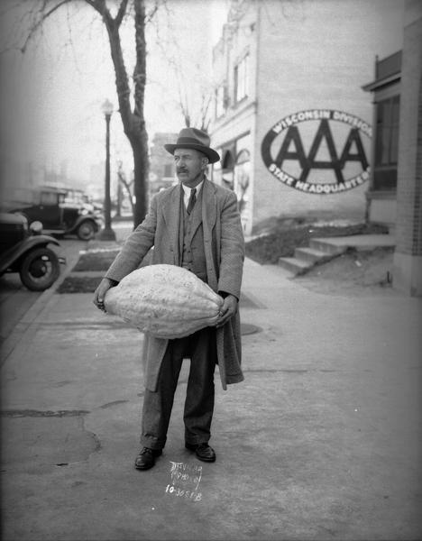 Albert A. Larson, Burke Township, displaying a 58 pound squash which he gave to the Madison Salvation Army. In the background is a sign on the side of a building for "AAA Wisconsin Division State Headquarters."