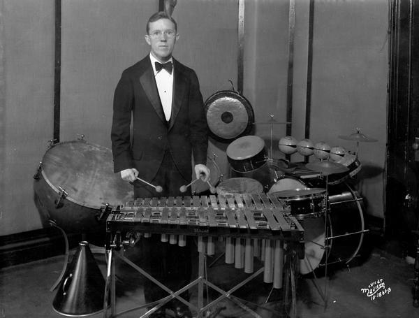 A tuxedo-clad percussionist standing at his marimbas, with other instruments including a gong, drum kit, and tympani behind him at the WIBA radio station.