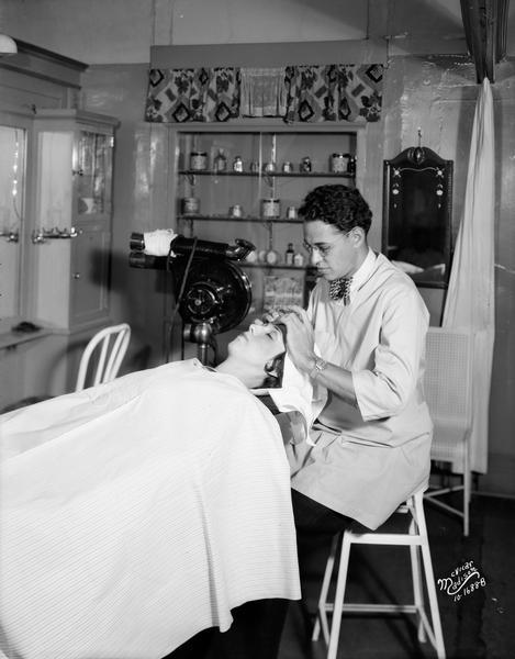 A male beauty operator is giving a woman a facial massage in the Baron Brothers Department Store Beauty Shop/"Bobber" Shop.