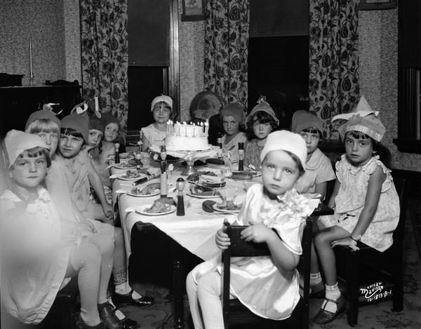 Eleven little girls in party hats sit around a dining table set with a birthday cake and candles, and party favors.