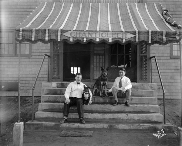 Chanticleer Orchestra managers, each with a dog at his side, sitting on the front steps of a building with an awning overhead that reads "Chanticleer."