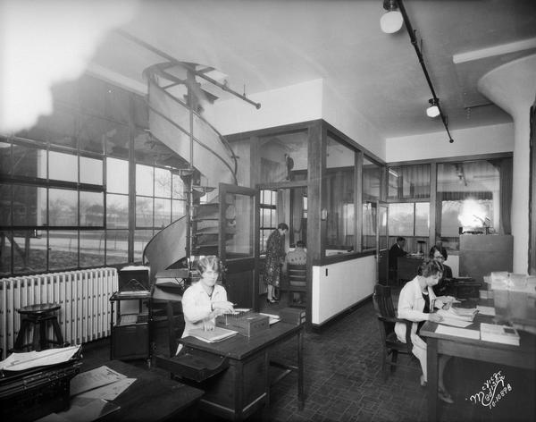 Office staff at work in Mautz Paint & Glass Company office, 939 East Washington Avenue. There is a spiral staircase on the left leading up to the next floor. Breese Stevens athletic field can be seen through the windows in the background.