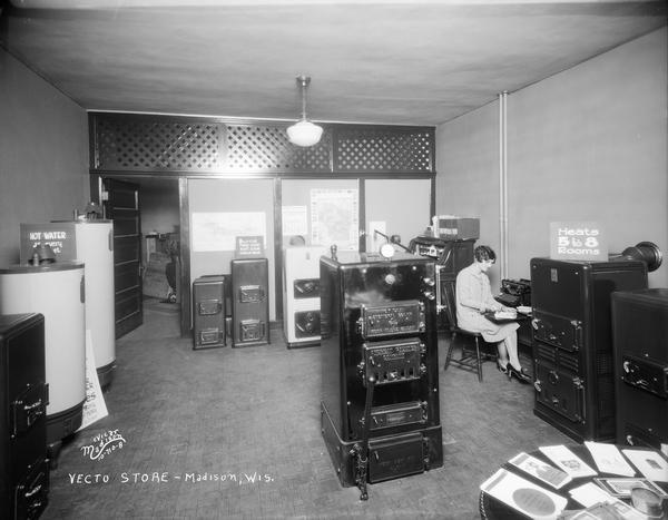 Interior of Vecto Store, located at 422 West Gilman Street. Included in the view are American Radiator Company furnaces, hot water heaters, and a woman sitting at a typewriter.