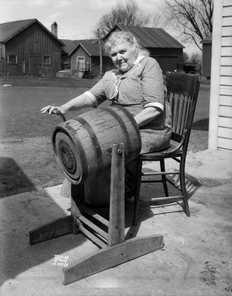 Ellen (Mrs. William) Lacy, of Sunny Slope farm, 5810 Lacy Road, south of Madison, sitting in the farmyard and turning a barrel butter churn.