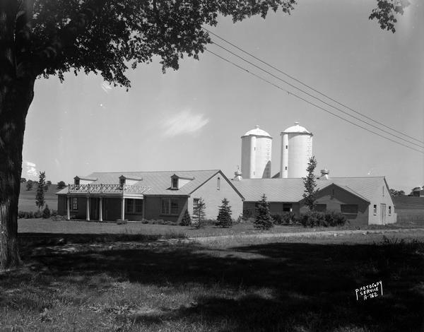 Rennebohm dairy farm buildings, showing house in foreground and barn and silos in background, framed by large tree, located 5 miles from the capitol on Highway 151 between Madison and Sun Prairie. The milk produced on the farm was used in the Rennebohm soda fountains.