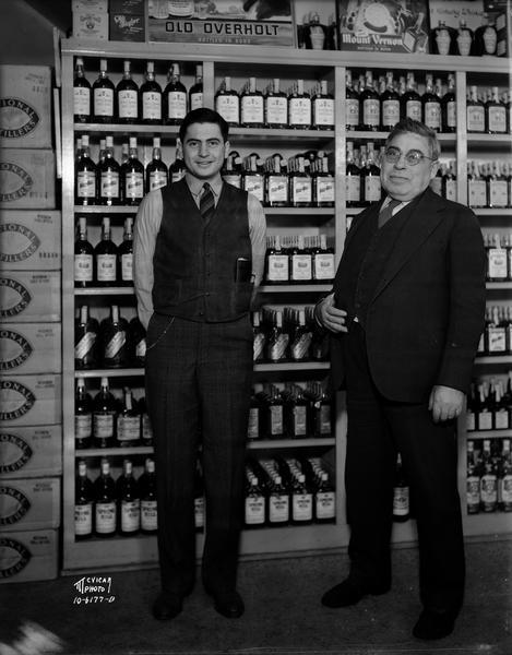 Saul Sinaiko and his son Donald Sinaiko, proprietors, standing in front of liquor bottle display, at the Badger Liquor Shop, located at 402 State Street.