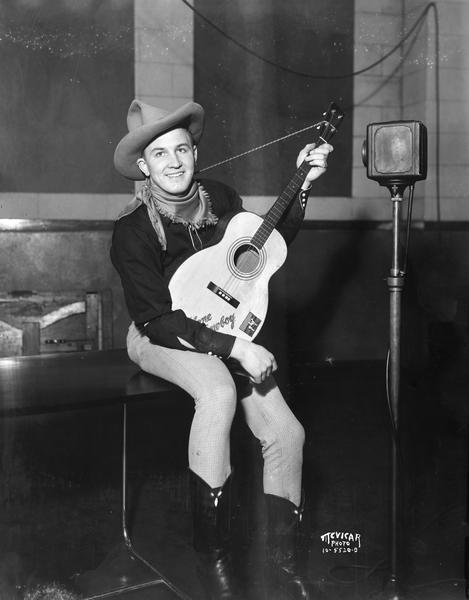A man dressed as a cowboy is sitting and holding a guitar in front of a microphone in W.I.B.A. studios, 111 King Street, Room 28.