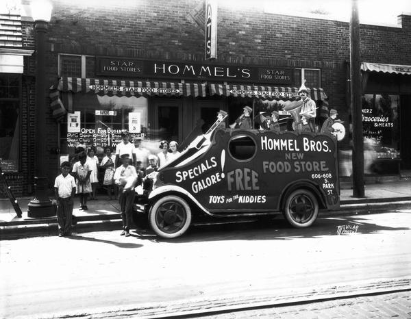 Hommel's Star Food Store at 606 S. Park Street, with promotional truck in front of store, and children looking on. Vehicle (Ballyhoo Truck) has a painted sign on the side: "Hommel Bros. new food store, specials galore! Free toys for the kiddies." There is a man dressed as a clown playing an instrument, and there are cardboard cutouts of other musicians.