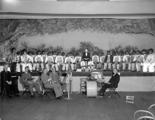 Minstrel show actors, sitting on stage, in costume and blackface with a small orchestra sitting below, at Luther Memorial Church, 1023 University Avenue.