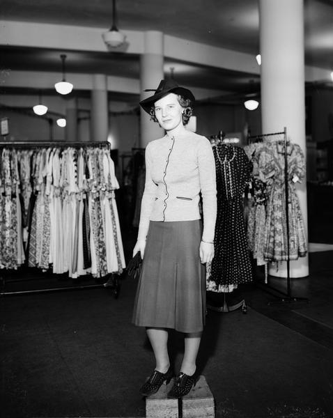 Model standing on box wearing jacket and skirt, hat, carrying gloves. Part of a fashion series from Kessenich's Ready to Wear, 201-203 State Street, with racks of dresses.