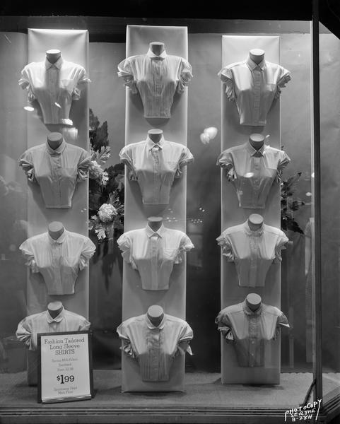Exterior night view night of Hill's Department Store window, 202 State Street, displaying twelve ladies' "fashion tailored" long sleeve shirts.  Advertised for sale for $1.99.