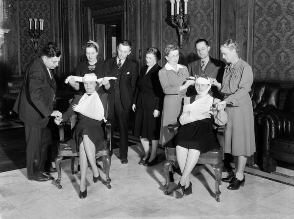 Red Cross first aid class of State government employees in the Wisconsin State Capitol Assembly parlor practicing bandaging - standing l to r: E.C. Giessel, Budget Bureau; Margaret Miller, instructor; W.C. Graham, Highway Commission; Catherine Collins, Tax Commission; Esther Schadde, instructor, Board of Health; G.M. Mathews, Banking Commission; and Marion Poole, instructor, Highway Commission. The victims are Audrey Hett (left) Bureau of Personnel and Ruth Heise, Board of Health.