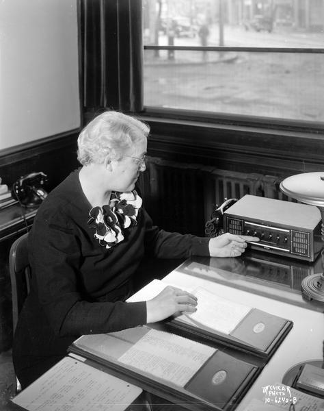 Regina Groves (Mrs. Earl Barnhart), sitting at her desk, operating an interdepartment communication system at the Groves School for Secretaries. Behind her is a window that looks out onto the street.