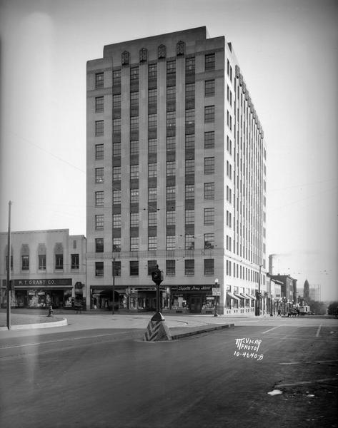 Vertical view of the Tenney Building, 110 East Main Street, taken from across the street. Visible are Liggett's Drug Store, Three Sisters and Mangel's women's clothing stores in the ground floor of the Tenney Building and the W.T. Grant store, 21-23 Pinckney Street. There is a traffic light and safety island for pedestrians in the middle of the street.