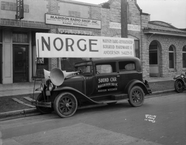 "Sound car" with loud speakers on the front hood, and a large sign on top that reads: "Norge, Madison Radio & Refrigerator Co., Borgrud Hardware Co., Anderson Sales Co." parked in front of the Madison Radio Shop, 1210 Regent Street.  Also shows Lumina Terrazzo & Tile Co., 1208 Regent Street.