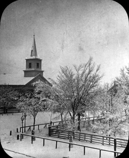 Elevated wintertime view of the intersection of South Carroll and West Main Streets, showing the First Baptist Church constructed in 1854, the wooden fence and hitching rail that surrounded the Capitol Square, and the street lights then in use. Before their building was constructed, the Baptists, like many other congregations, worshiped in the Wisconsin State Capitol building. The State Historical Society rented space in the basement of the Baptist Church until 1866 when it moved across the street to the Capitol. The wooden fence in this photograph was replaced with an ornamental iron and stone fence in 1873.
