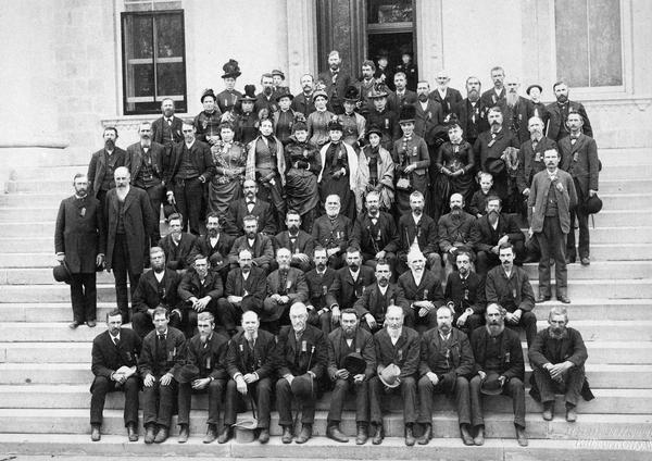 Group portrait of the 16th Wisconsin Infantry at the Wisconsin State Capitol, together with their wives and families, taken at their first reunion. Because this picture was photographed by H.H. Bennett, a veteran of the 12th Wisconsin Infantry, some members of that regiment may also be present. The veterans are thought to be posing on the steps of the South Wing of the Capitol (as opposed to the identical North Wing), because the fourth floor of the north addition was entirely given over to a large convention room.