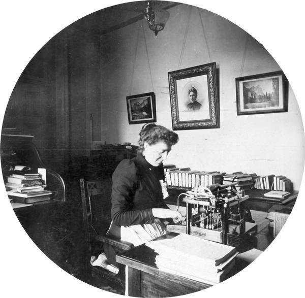 Minnie Oakley, assistant librarian of the Wisconsin Historical Society, seated at her desk in the South Wing of the Wisconsin State Capitol.  This photograph, combined with a similar portrait of Emma Hawley, suggests that librarians included aprons in their work wardrobes.  This round portrait was taken by Reuben Gold Thwaites, secretary of the Historical Society and an avid amateur photographer, using the popular Kodak 2 format.
