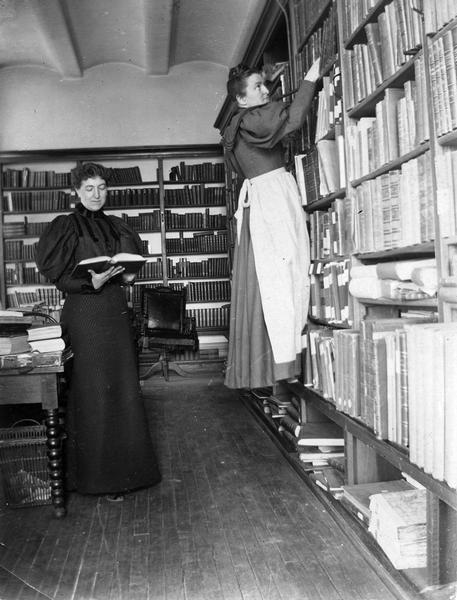 Minnie Oakley and Florence Baker Hayes (reaching for a book), two Historical Society librarians, during the period when the Society was housed in the South Wing of the Wisconsin State Capitol. Details of the Capitol furnishing and construction in this photograph include the leather swivel chair (in need of repair) and the Jenny Lind table, as well as documentation of the ceiling construction.